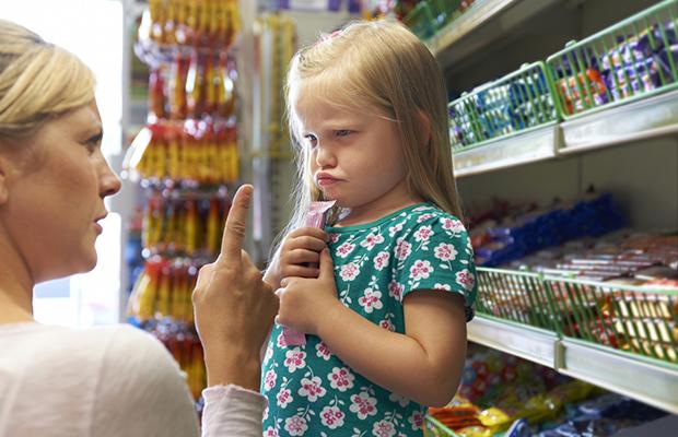 kneeling mother holding finger up to little, pouting girl in front of candy shelves in a store